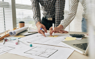 A team of architects meeting to discuss details of architectural projects. On the table, there are floor plans, rulebooks, markers, a laptop, coffee, and a calculator.
