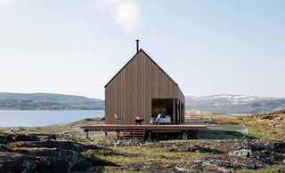 Exterior design of a single-story cabin elevated on pier foundations next to a river, showcasing the deck area and a glimpse of the interior space through an open window. The surroundings feature a flat valley with green and orange vegetation