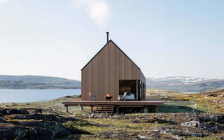 Exterior design of a single-story cabin elevated on pier foundations next to a river, showcasing the deck area and a glimpse of the interior space through an open window. The surroundings feature a flat valley with green and orange vegetation