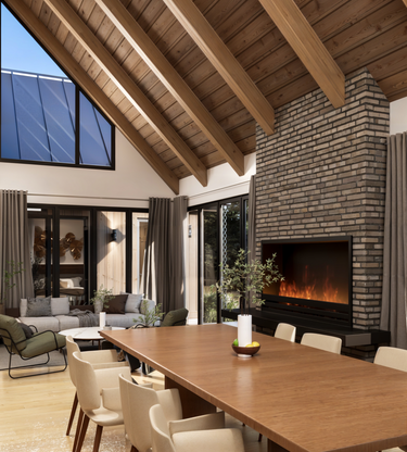A dining area with a vaulted ceiling, seamlessly connected to the kitchen and living space in a cabin home design. Featuring a central stone chimney and tall open windows that provide views of the outdoors.