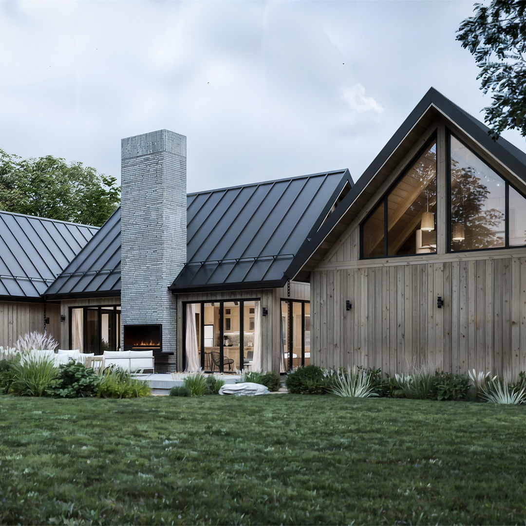 A courtyard view of a modern cabin showcasing a tall brick chimney as the central feature. The building has black metal roofs and natural wood siding. Glass doors with curtains flank the chimney, leading to an interior living space. The outdoor area includes a patio with seating, surrounded by landscaped greenery and ornamental grasses. The bright blue sky enhances the clean, minimalist aesthetic of the design.