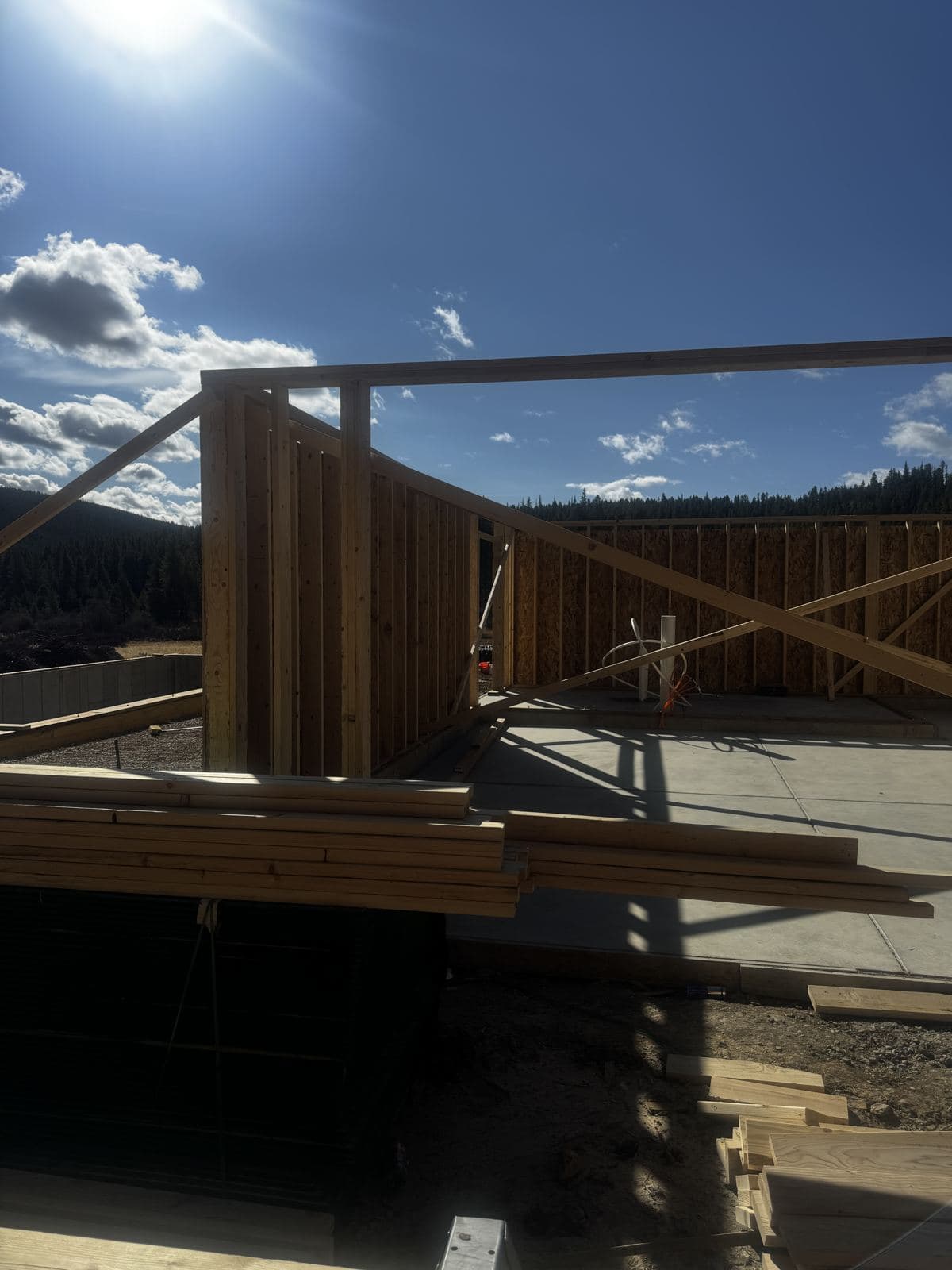 Stud walls and bracing on the slab, lumber stacked in front, the ridge behind.