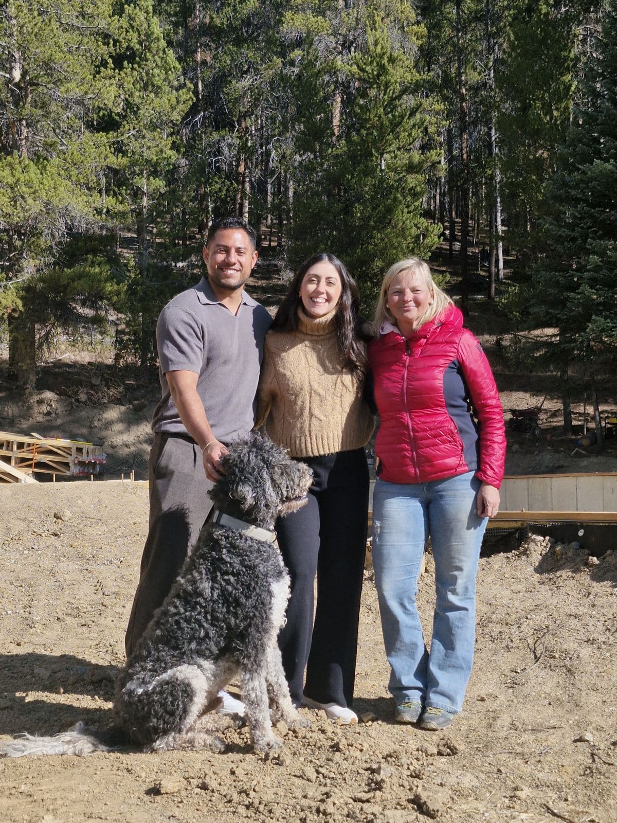 Three people and a dog on the site, pines behind.