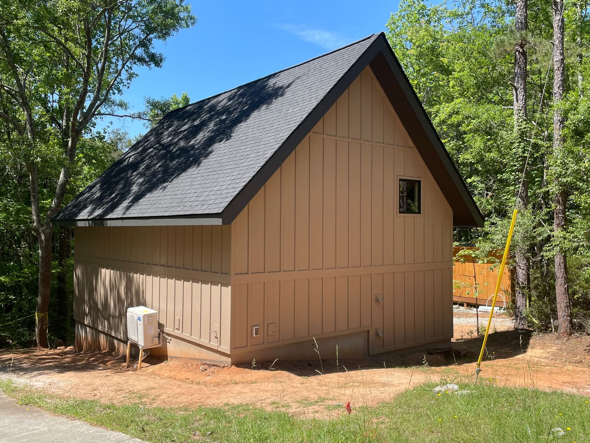 A tan board-and-batten gable with a dark shingle roof, set in pines on red soil.