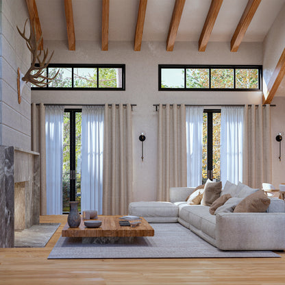 Rustic modern living room with high ceilings, exposed wooden beams, and large black-framed windows. Features a cozy beige sectional sofa with neutral throw pillows, a reclaimed wood coffee table, and a stone fireplace with a mounted antler decor. Natural light enhances the warm, inviting ambiance, complemented by soft linen curtains and elegant black wall sconces. Perfect blend of contemporary and cabin-style interior design