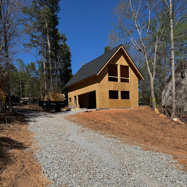 Wooden cabin under construction with a gravel driveway in a forested area