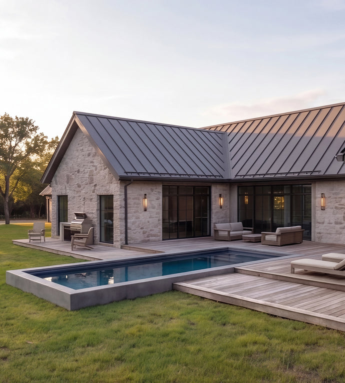 The Cedar Residence - Modern single-story house with a stone exterior, dark metal roof, large glass windows, a concrete hot tub, and a wooden deck with outdoor seating, set on a grassy lawn at dusk.