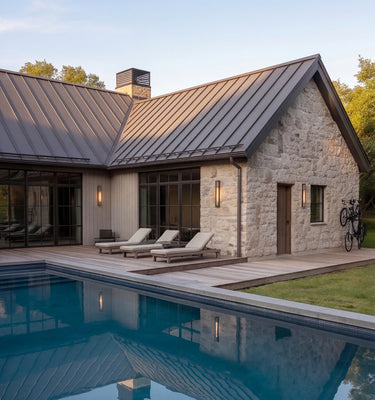 The Cedar Residence - Modern single-story house with a stone exterior, dark metal roof, large glass windows, a wooden deck, three outdoor lounge chairs, and a swimming pool in the foreground.