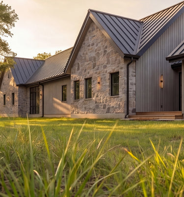 The Cedar Residence - Modern farmhouse-style house exterior with stone and wood siding, dark metal roofs, and a grassy lawn in the foreground at sunset.