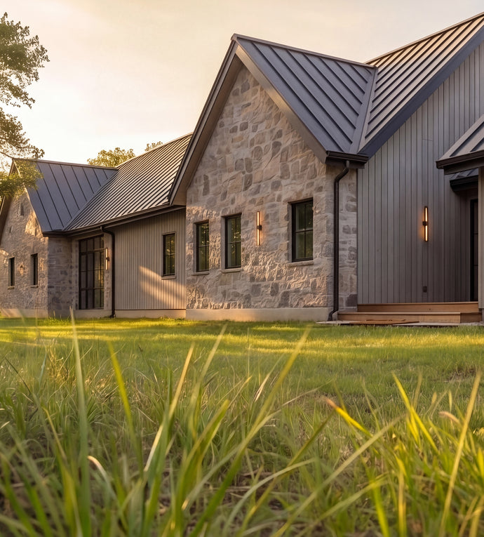 The Cedar Residence - Modern farmhouse-style house exterior with stone and wood siding, dark metal roofs, and a grassy lawn in the foreground at sunset.