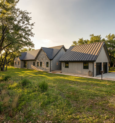 The Cedar Residence - A modern rustic cabin complex with gray siding and dark metal roofs, set in a grassy field with trees under a warm sky.