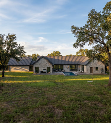 The Cedar Residence - A modern single-story ranch-style home with a stone exterior, dark metal roof, large glass windows, and a pool, shown in a grassy outdoor setting with trees under a blue sky.