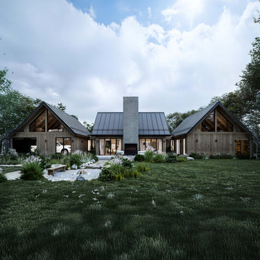 Modern farmhouse-style home with three gabled sections connected by a central structure featuring a tall stone chimney and metal roof. The exterior is clad in wood siding, and large windows reveal glimpses of the interior. The landscaped backyard includes a stone pathway, lush greenery, ornamental grasses, and a small seating area.