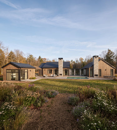 Coyotes Den: A large modern farmhouse-style house with wood siding, dark metal roofs, stone chimneys, and a backyard pool, shown in a landscaped yard with a gravel path and wildflowers under a blue sky.