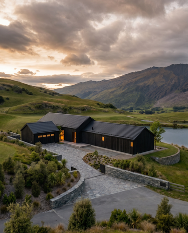 Casa del Prado - A modern black wooden cabin with a matching small outbuilding, set in a scenic mountain valley at sunset with rolling green hills and a stone driveway.