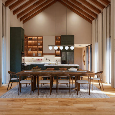 A spacious open-concept kitchen and dining area with a vaulted ceiling featuring exposed wooden beams. The kitchen has dark green cabinetry, warm wood shelving with integrated lighting, and a large marble island with barstools. A wooden dining table with eight matching chairs sits on a textured area rug, illuminated by a modern pendant light with globe fixtures. Floor-to-ceiling curtains frame large windows, allowing natural light to enhance the warm, earthy tones of the space