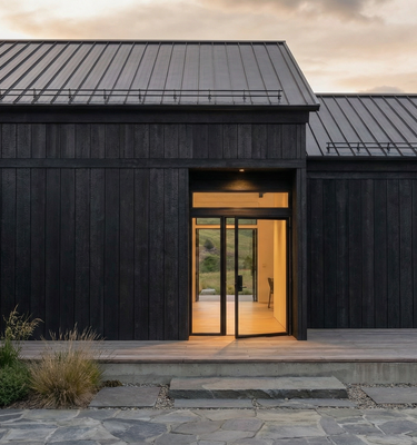 Casa del Prado - Modern black wood-paneled house exterior with a dark metal roof, double glass entry doors, warm interior lighting, stone steps, and a stone driveway at dusk.