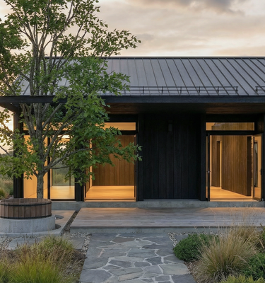 Casa del Prado - Modern single-story house exterior with dark wood and metal roofing, large glass doors, warm interior lighting, a stone walkway, landscaped grasses, and a tree in a rural outdoor setting at sunset.