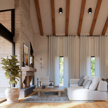 A spacious living room with vaulted ceilings featuring exposed wooden beams, a stone fireplace, and floor-to-ceiling curtains. The space is decorated with a neutral-toned sectional sofa, a rustic wooden coffee table, and a cozy armchair, blending modern and rustic design elements.