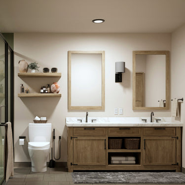 A modern rustic bathroom featuring a double vanity with a natural wood finish, a white marble countertop, and two framed mirrors. The vanity includes black metal hardware, open shelving with woven baskets, and neatly folded towels. To the left, floating wooden shelves hold decorative vases, toiletries, and essentials. A sleek white toilet sits below the shelves, with a black toilet brush and paper holder. A soft gray rug lies on the floor, complementing the neutral color palette. The space is softly lit by 