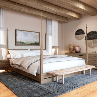 A cozy bedroom featuring a wooden four-poster bed with soft neutral bedding, a textured area rug, and rustic decor. The space has exposed wooden ceiling beams, warm lighting, and a mix of modern and bohemian design elements with earthy tones.
