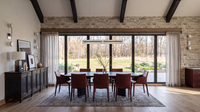 The Maine Project: A modern dining room featuring a dark wood dining table and six reddish-brown upholstered chairs on a patterned area rug, set against a stone wall and large floor-to-ceiling windows.