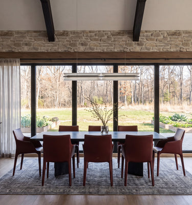The Maine Project: A modern dining room featuring a dark wood dining table and six reddish-brown upholstered chairs on a patterned area rug, set against a stone wall and large floor-to-ceiling windows.
