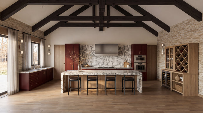 The Maine Project: A high-ceilinged kitchen interior with exposed dark wood beams, stone walls, a marble island, bar stools, cabinetry, and a wine rack.