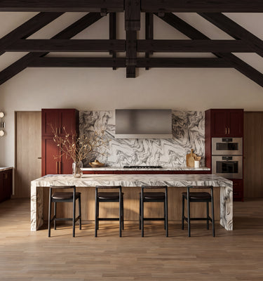 The Maine Project: A high-ceilinged kitchen interior with exposed dark wood beams, stone walls, a marble island, bar stools, cabinetry, and a wine rack.