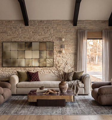 The Maine Project: A styled living room interior featuring a cream sofa, two brown armchairs, a wooden coffee table, a patterned area rug, a stone brick wall, and a large rectangular wall art piece.