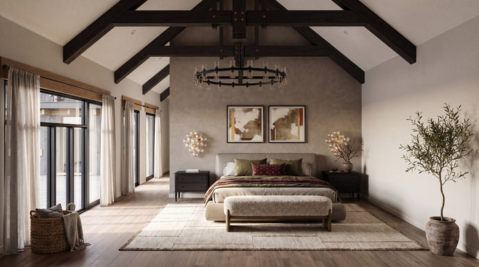 The Maine Project: A styled bedroom interior with a bed centered against a textured plaster wall, flanked by dark wood nightstands and framed art. The room features a high vaulted ceiling with exposed dark wood beams, a large rug, wood flooring, and a potted tree on the right.