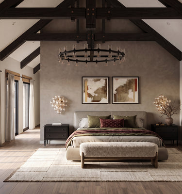 The Maine Project: A styled bedroom interior with a bed centered against a textured plaster wall, flanked by dark wood nightstands and framed art. The room features a high vaulted ceiling with exposed dark wood beams, a large rug, wood flooring, and a potted tree on the right.