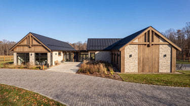 The Maine Project: A modern farmhouse-style house with large wood-paneled gable roofs, stone walls, a dark metal roof, and a paved driveway in an outdoor setting under a clear blue sky. 