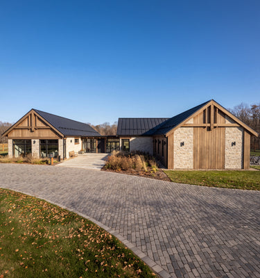 The Maine Project: A modern farmhouse-style house with wood and stone exterior walls, dark metal roofs, and a curved gray paved driveway on a grassy lawn under a clear blue sky.