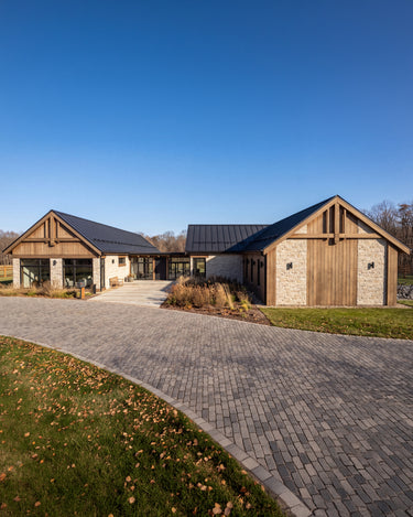 The Maine Project: A modern farmhouse-style house with wood and stone exterior walls, dark metal roofs, and a curved gray paved driveway on a grassy lawn under a clear blue sky.