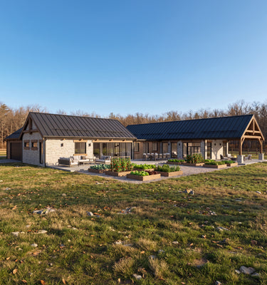 The Maine Project: A modern single-story house with a dark metal roof, light stone exterior, and large glass walls, shown on a grassy lot under a clear blue sky.