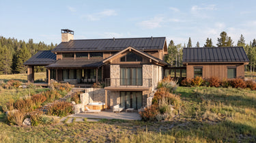 The Hunters Lodge: A large modern mountain-style lodge with dark metal roofing, wood siding, and stone walls, shown on a grassy hillside with trees in the background.