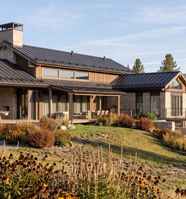 The Hunters Lodge: A large modern mountain-style house with dark metal roofing, wood siding, and stone walls, situated on a grassy hillside with trees and shrubs in the foreground under a blue sky.