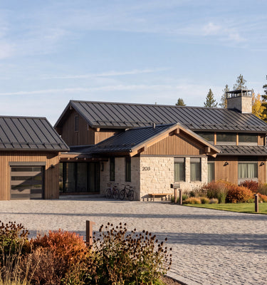 The Hunters Lodge: A modern single-story log cabin style home with dark metal roofs, horizontal wood siding, and a stone facade, set on a gravel driveway with landscaped landscaping and trees in the background.