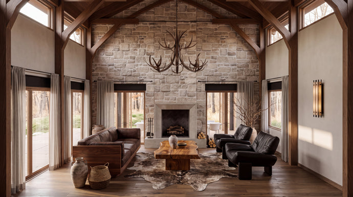 The Hunters Lodge:  A rustic, high-ceilinged lodge-style living room with a stone fireplace, exposed wood beams, large windows, a brown leather sofa, a black leather armchair, a wooden coffee table, and a large antler chandelier.