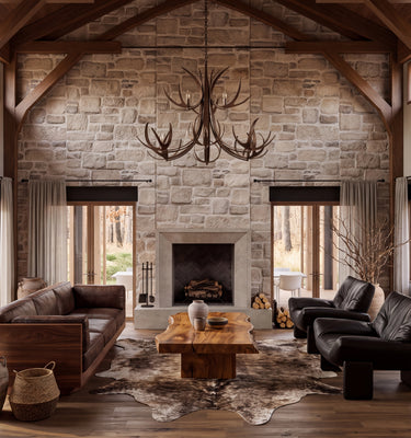 The Hunters Lodge:  A rustic, high-ceilinged lodge-style living room with a stone fireplace, exposed wood beams, large windows, a brown leather sofa, a black leather armchair, a wooden coffee table, and a large antler chandelier.
