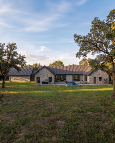 The Cedar Residence - A modern single-story ranch-style home with a stone exterior, dark metal roof, large glass windows, and a small pool, set on a grassy lot with trees under a blue sky.