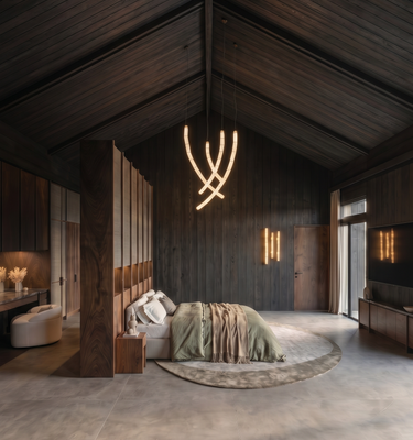 Casa del Prado - A luxury modern bedroom featuring a large bed with sage green and beige bedding, set in a spacious room with dark wood-paneled walls and ceiling, a circular concrete floor, and a sculptural illuminated pendant light.