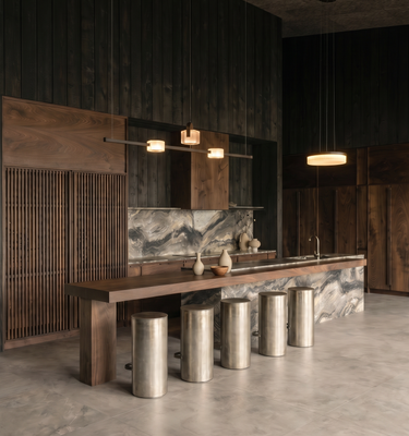 Casa del Prado - A modern interior space with a long wooden bar or counter set over a marble-topped island, supported by six cylindrical metallic stools. The room features dark wood-paneled walls, a concrete floor, pendant lights, and a marble backsplash.